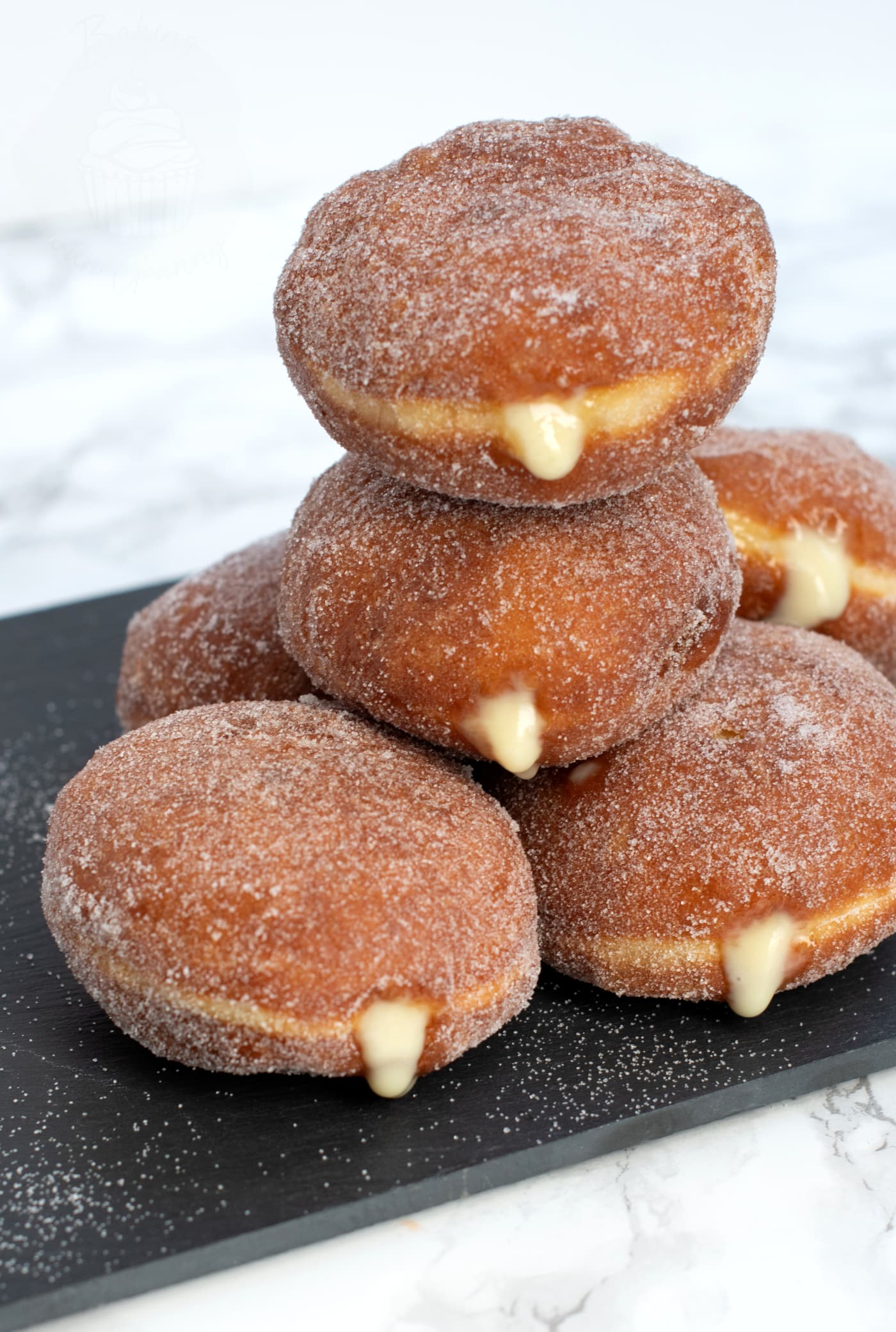 Stack of custard donuts on a black slate chopping board. The custard can be seen oozing from the sides of the sugar coated donuts.