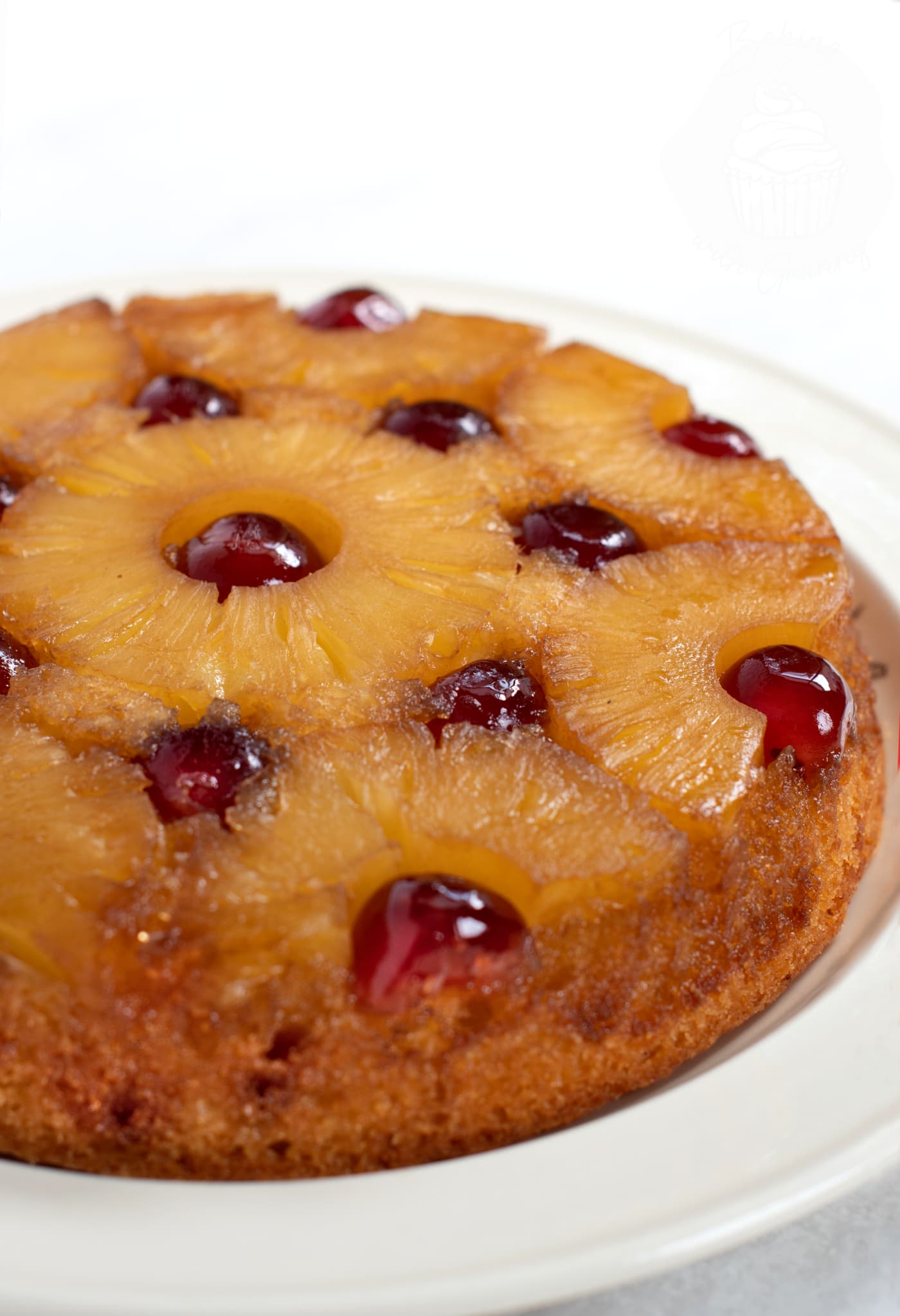 Pineapple Upside Down Cake on a light cream plate and white background. The red cherries are reflecting the light and the pineapple rings are golden yellow.
