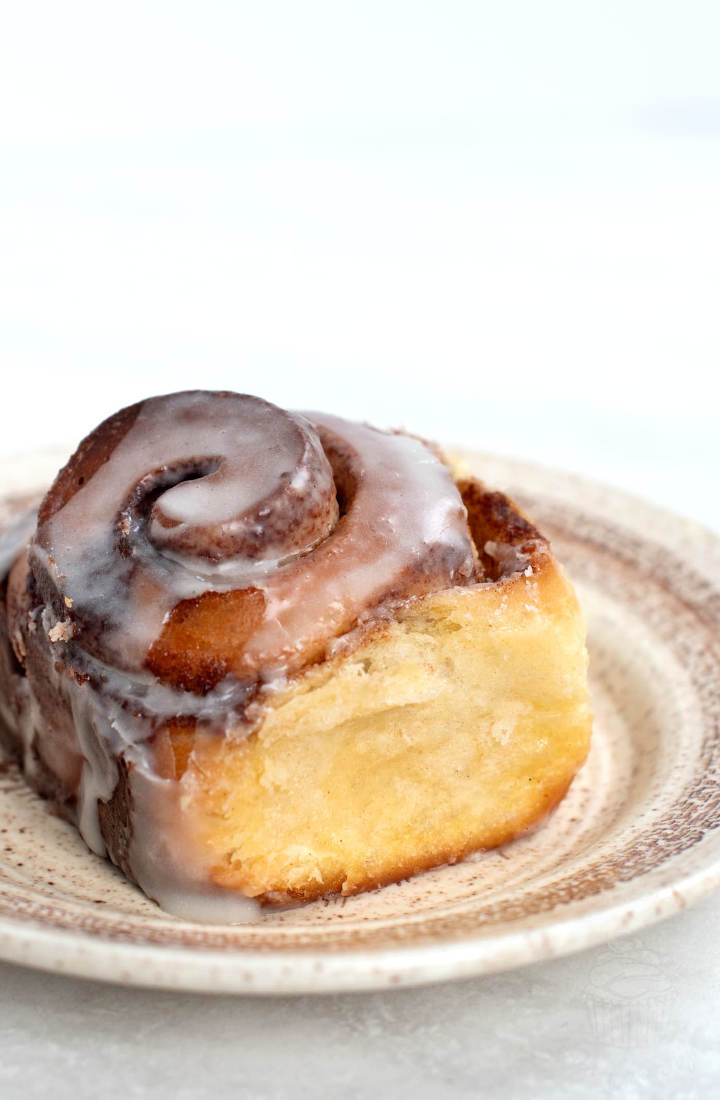 A close-up of a frosted cinnamon bun on a beige plate. The roll is golden brown with visible swirls and topped with a glossy layer of icing, reminiscent of an authentic UK recipe. The background is a light, soft-focus setting.