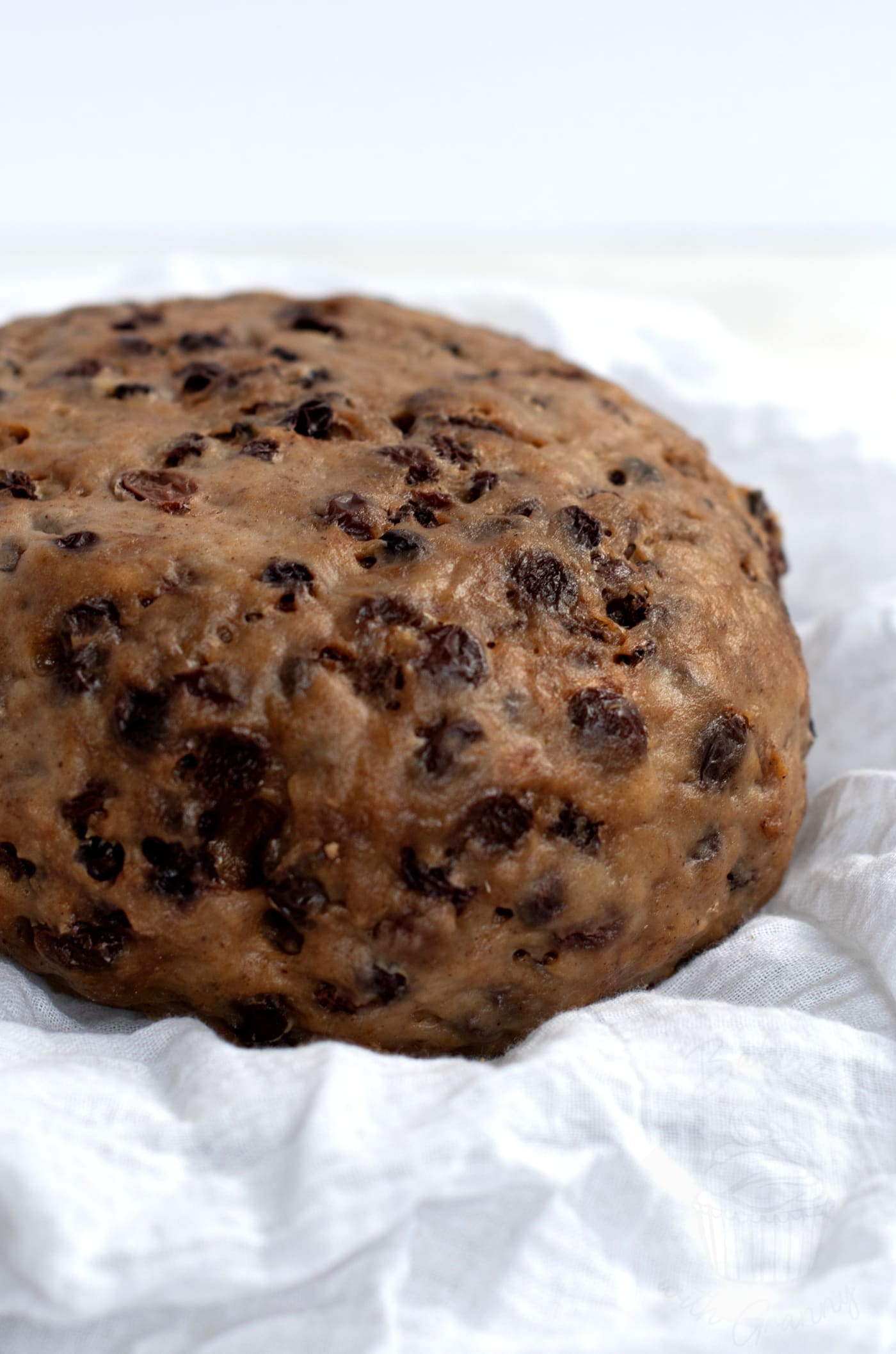 A round, dense traditional Scottish clootie dumpling, filled with visible raisins and currants, rests on a white cloth. The dumplings textured surface reflects its rich, moist composition.