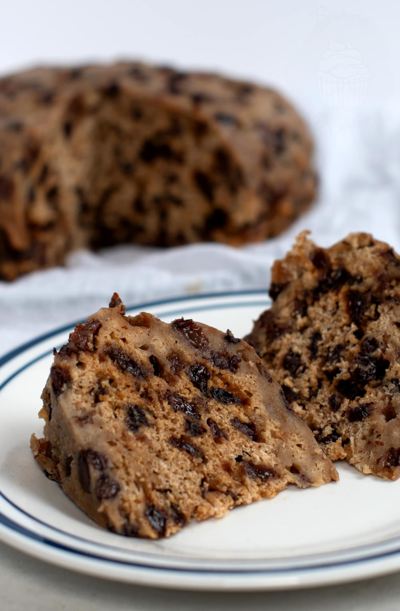 A close-up of two slices of traditional Scottish clottie dumpling, rests on a white plate with blue trim. The clootie dumpling, filled with dark fruit pieces, appears moist and dense. A larger portion is blurred in the background, nestled on white cloot.