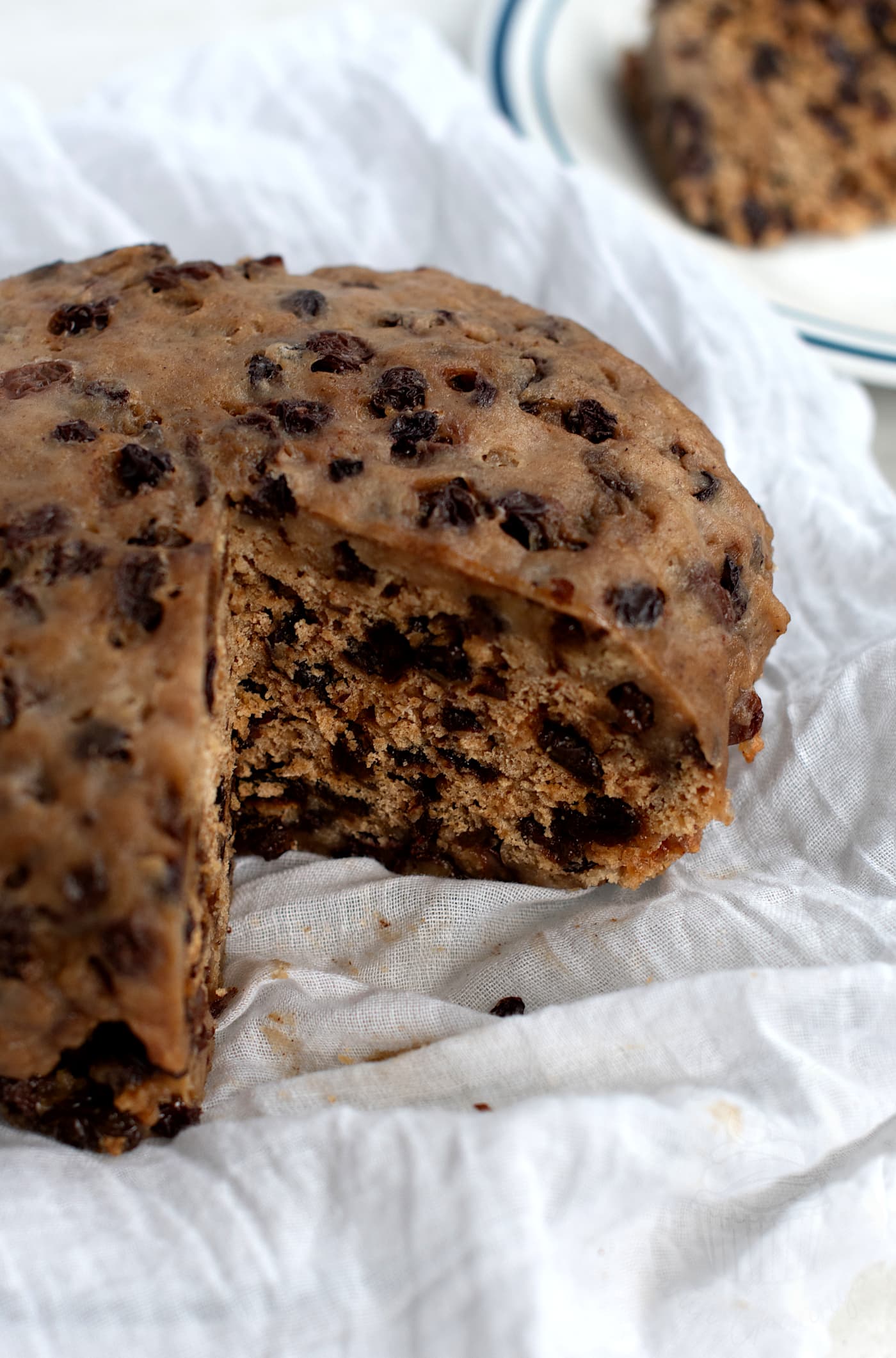 A close-up of a Scottish duff on a white cloth evokes the charm of a traditional clootie dumpling recipe. The dumpling is sliced to reveal its dense texture filled with dark raisins and dried fruits. A small portion rests on a white plate in the background, inviting a taste of nostalgia.