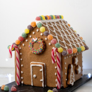 A gingerbread house crafted from a delightful UK recipe is adorned with colourful jelly tots, striped candy canes, and white icing patterns. A wreath made of sprinkles graces the front. The house rests on a black slate surrounded by scattered sweeties.