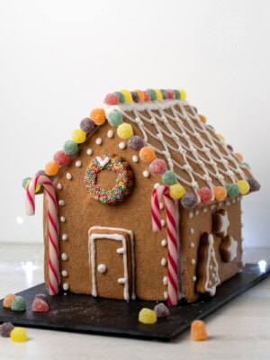 A gingerbread house crafted from a delightful UK recipe is adorned with colourful jelly tots, striped candy canes, and white icing patterns. A wreath made of sprinkles graces the front. The house rests on a black slate surrounded by scattered sweeties.