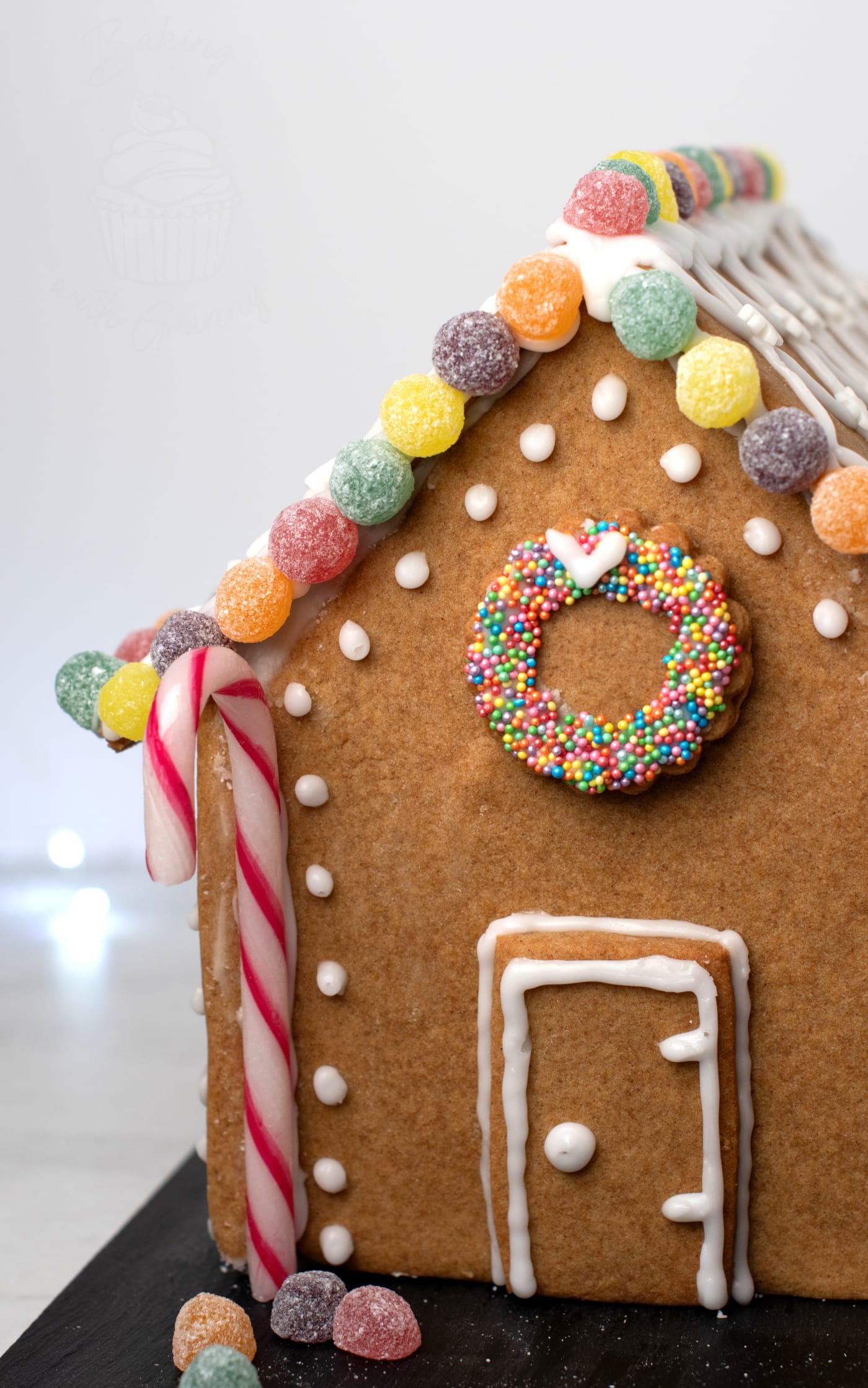 Front detail of a gingerbread house with piped icing door, candy cane columns and a sprinkle-covered biscuit wreath.