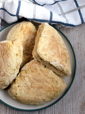 A plate of freshly baked, triangular shaped traditional Irish soda farls, sits on a wooden table. A striped white and blue kitchen towel is placed nearby.