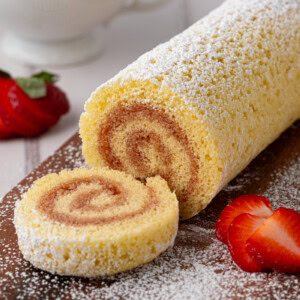 A classic Swiss Roll cake dusted with icing sugar elegantly sliced on a wooden board. Beside it, a cup of tea and fresh strawberry slices evoke UK charm. The golden cake reveals a mesmerizing spiral pattern, inviting you to try this delightful recipe.