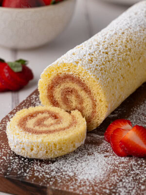 A rolled sponge cake, reminiscent of a classic Swiss Roll recipe, is dusted with icing sugar and sliced on a wooden board. Fresh strawberries are placed around it, with a bowl of strawberries in the background.