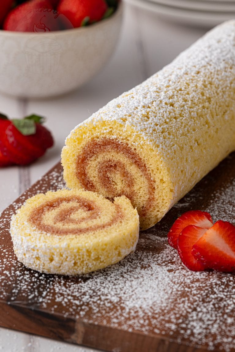 A rolled sponge cake, reminiscent of a classic Swiss Roll recipe, is dusted with icing sugar and sliced on a wooden board. Fresh strawberries are placed around it, with a bowl of strawberries in the background.