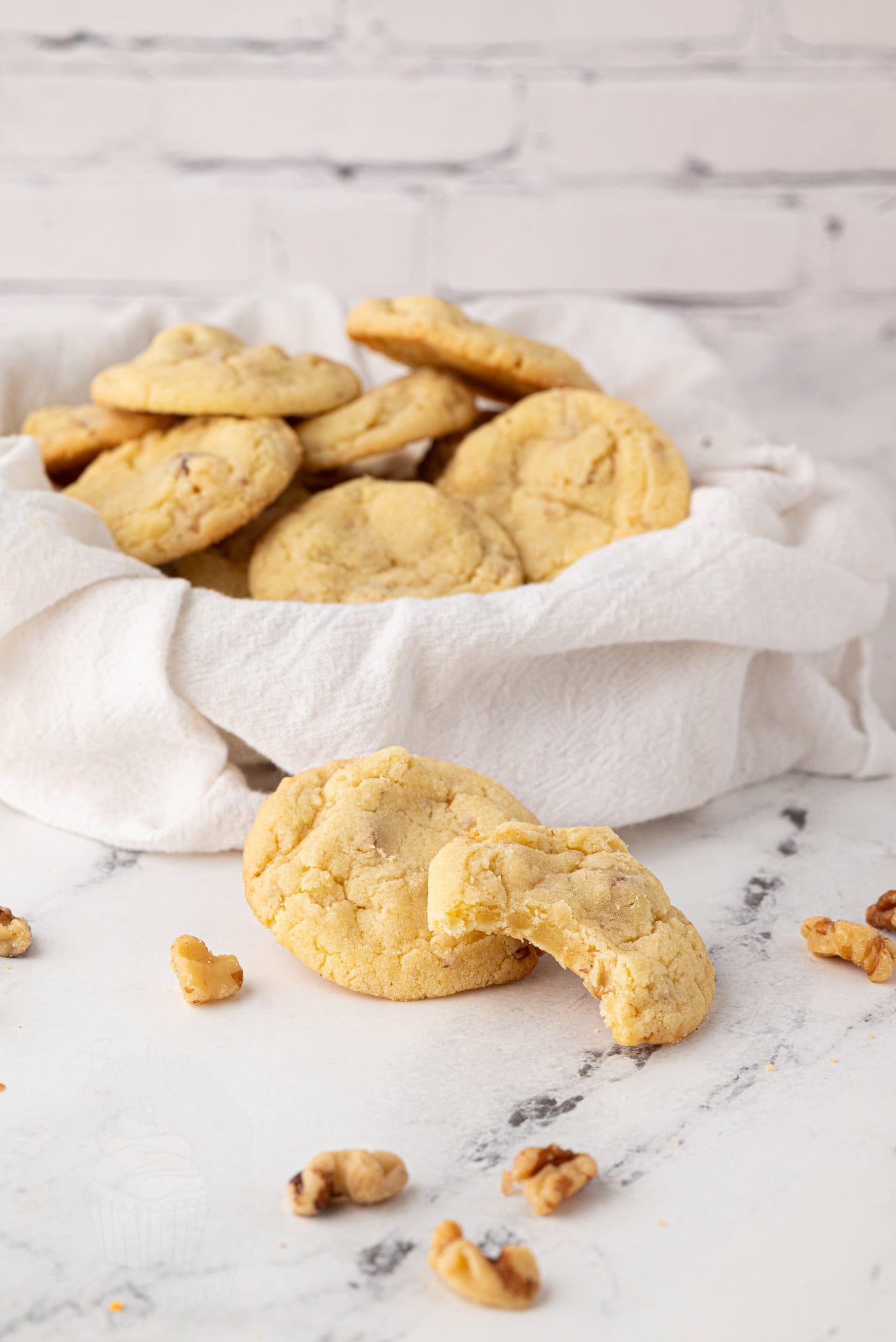 A basket filled with freshly baked walnut cookies on a white cloth. A bitten cookie and walnuts are scattered on a marble surface in front. The background features a white brick wall.