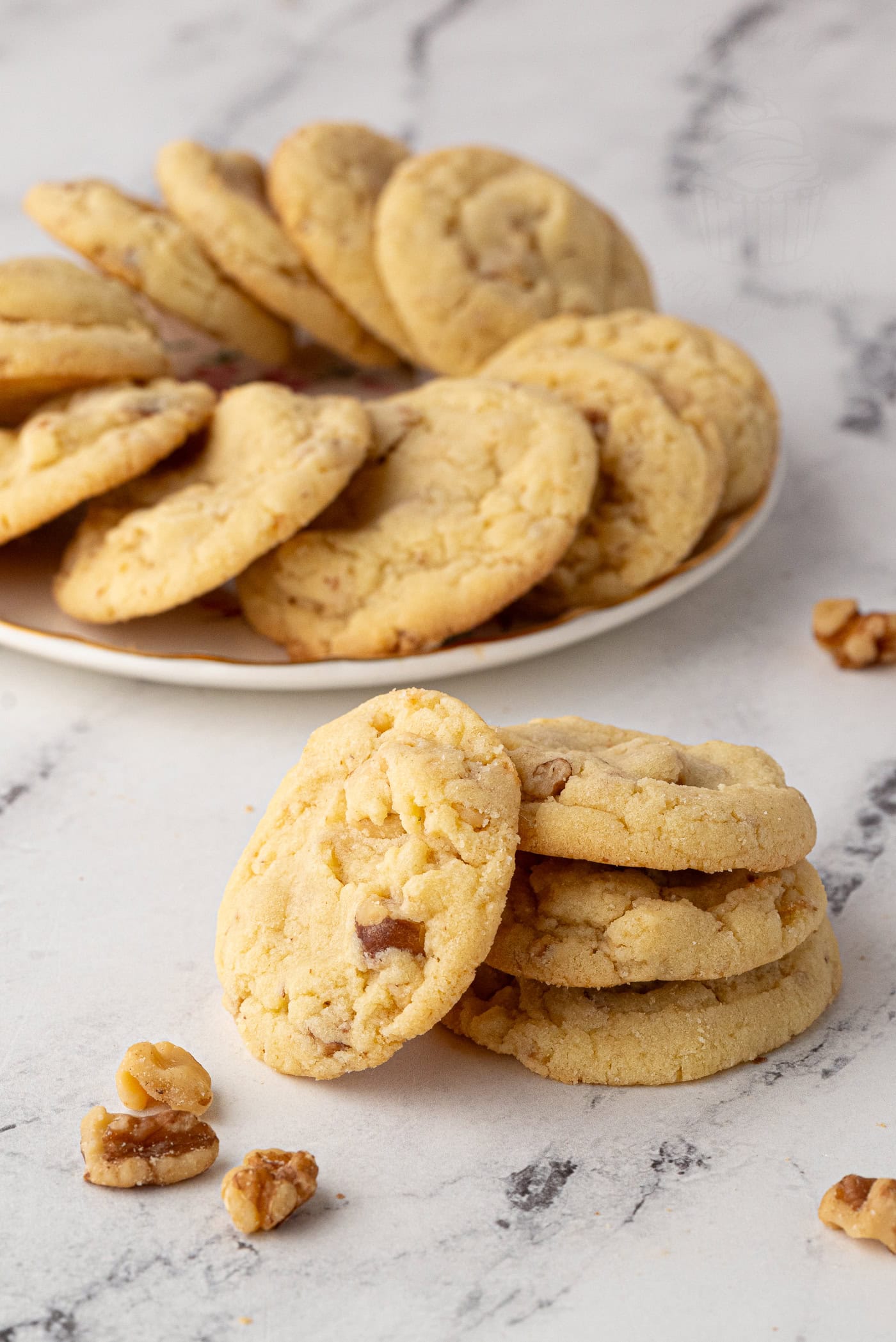 A stack of homemade walnut cookies filled with chunks of nuts. In the background, more cookies are piled on a white plate. They are arranged on a marble surface, with a few nuts scattered nearby.