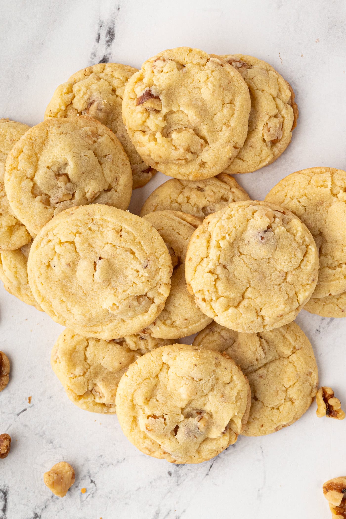 A pile of freshly baked walnut biscuits rests on a marble surface, with a few walnut pieces scattered around.