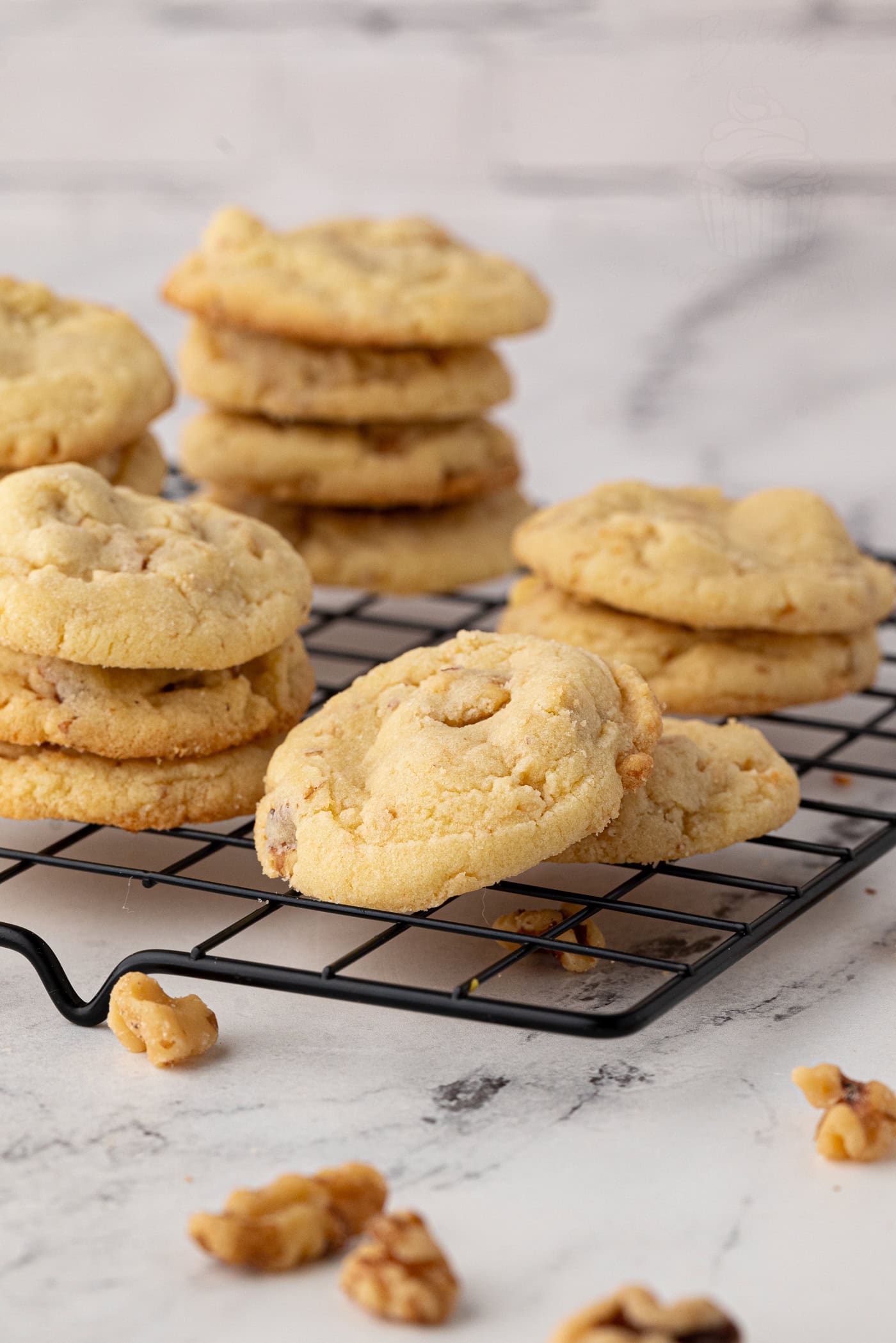 A cooling rack holds stacks of walnut cookies, fresh from the oven, with walnuts scattered around. The golden, textured treats rest on a white marble surface. This scene offers a glimpse into a warm and inviting baking atmosphere, perfect for trying out your favourite walnut cookies recipe.