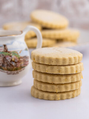 A stack of round, scalloped-edge traditional Scottish Tantallon Cake biscuits sits next to a decorative jug with a landscape design of North Berwick. More cookies are blurred in the background, all on a light surface, evoking the charm of an age-old recipe.