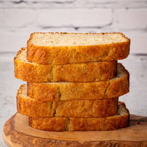 A stack of five thick slices of banana loaf bread rests on a wooden cutting board, hinting at the perfect recipe. The background features a crisp white brick wall, adding charm to this cosy kitchen scene.