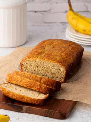 A loaf of banana loaf cake with three slices cut and placed on a wooden board. Ripe bananas, a white canister, and a stack of plates are in the background, all set against a light-coloured brick wall.