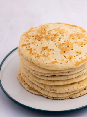 A stack of eight bubbly Scottish crumpets with a light golden-brown colour, features small holes on the surface and sits on a white plate with a green rim. The background is a soft, blurred white.