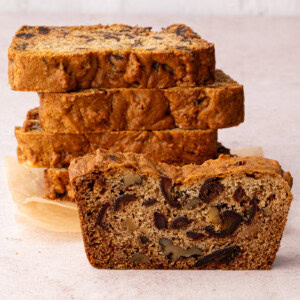A sliced date and walnut loaf reveals its moist, dense interior, adorned with visible walnuts and dates on a light surface. In the background, several golden-brown slices are stacked with parchment paper, showcasing an irresistible take on a classic recipe.