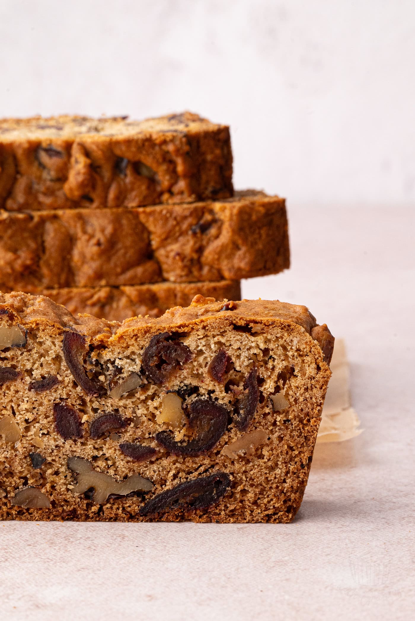 Slices of date and walnut cake are stacked on a light surface. The loaf cake has a golden-brown crust with visible chunks of nuts and fruit inside, resembling a traditional date walnut loaf. The background is softly blurred, highlighting the breads texture.