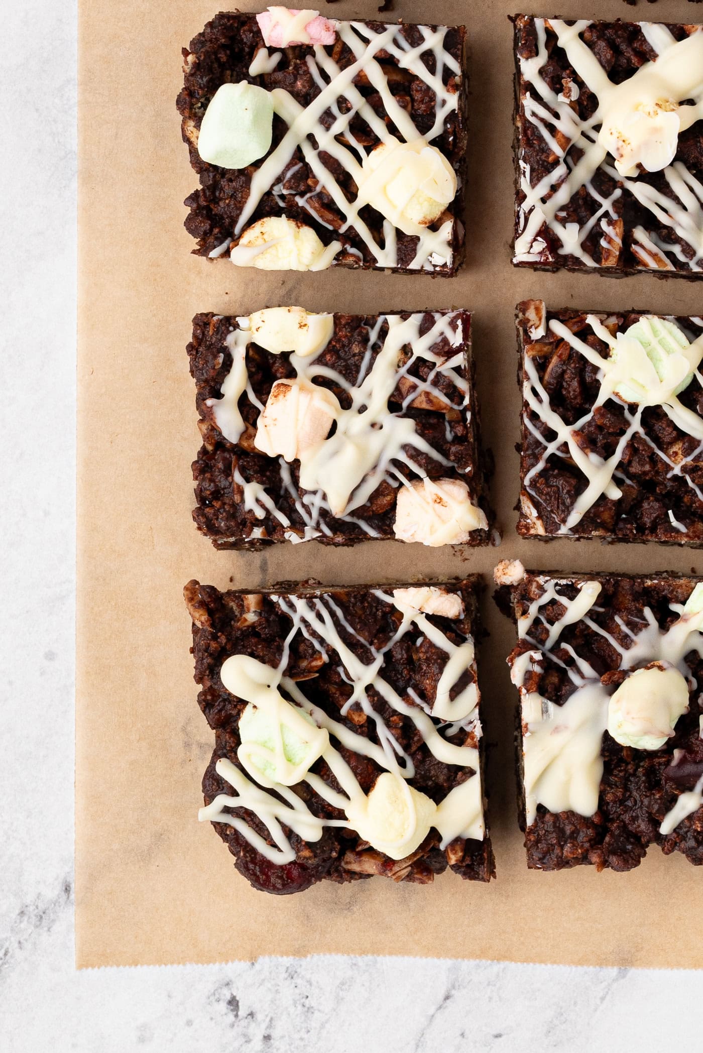 Six square pieces of chocolate rocky road, are topped with colourful marshmallows and drizzled with white chocolate. They sit invitingly on brown parchment paper against a marble surface.