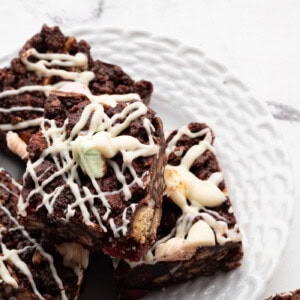 A plate of rocky road bars from a beloved rocky road recipe is topped with white chocolate drizzle. Filled with chocolate, marshmallows, cherries and nuts, these treats rest on a textured white plate against a marble surface.