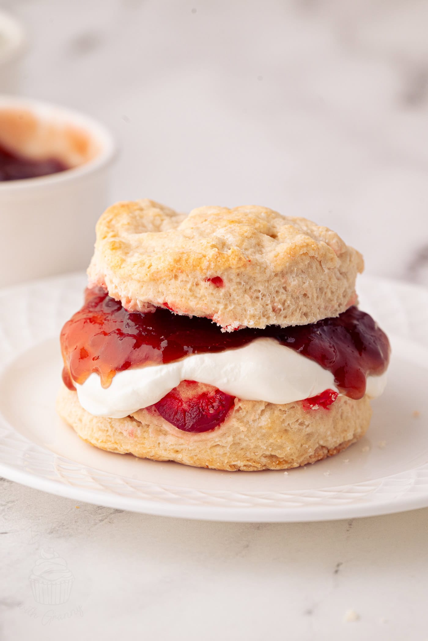 A cherry scone sandwich on a white plate, filled with fresh cream and strawberry jam. The golden-brown, crumbly scone pairs perfectly with the glossy jam. Its set against a light marble surface, inviting you to enjoy this delightful treat inspired by a beloved cherry scone recipe.