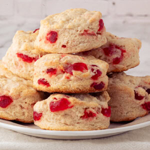 A plate of freshly baked cherry scones featuring bits of bright red glace cherries, stacked in a pyramid shape. The scones have a golden-brown crust and are set against a white brick background, offering an irresistible cherry scones recipe visual delight.