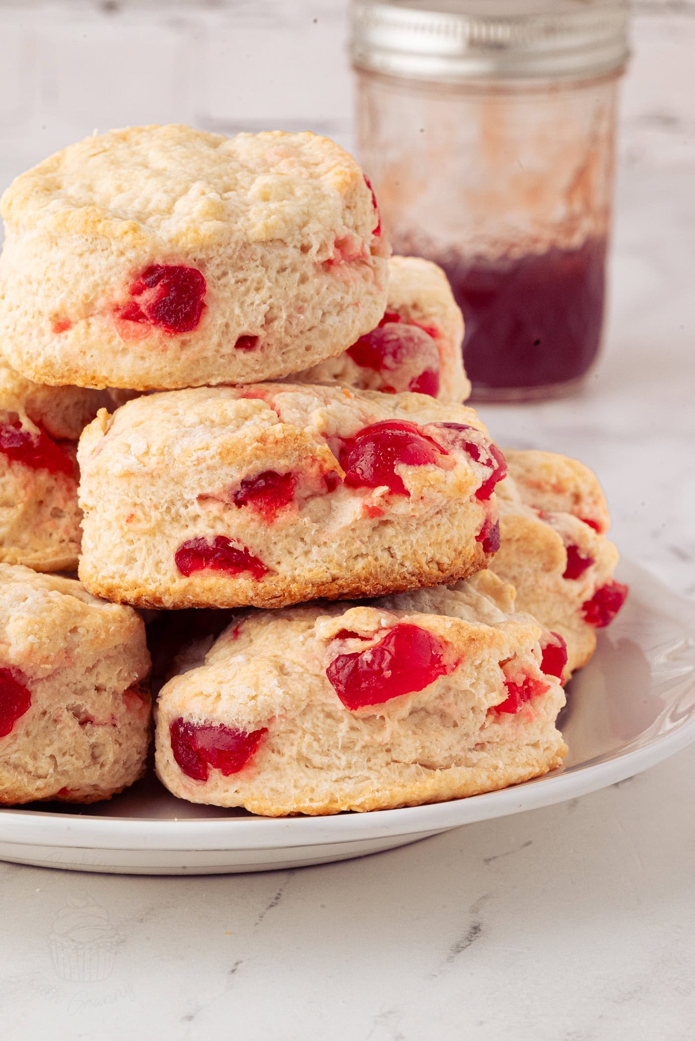 A delightful cherry scone recipe comes alive with visible chunks of red glace cherries. The scones, artfully stacked on a white plate, are paired perfectly with a jar of red jam in the background on a marble surface.
