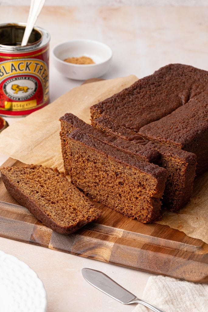 Sliced gingerbread loaf rests on a wooden cutting board, its deep brown hues inviting. Nearby, a can of black treacle and a small white dish with brown sugar add to the scene, accompanied by a knife and plate. The loaf is carefully placed on parchment paper.