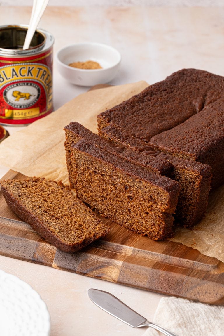Sliced gingerbread loaf rests on a wooden cutting board, its deep brown hues inviting. Nearby, a can of black treacle and a small white dish with brown sugar add to the scene, accompanied by a knife and plate. The loaf is carefully placed on parchment paper.