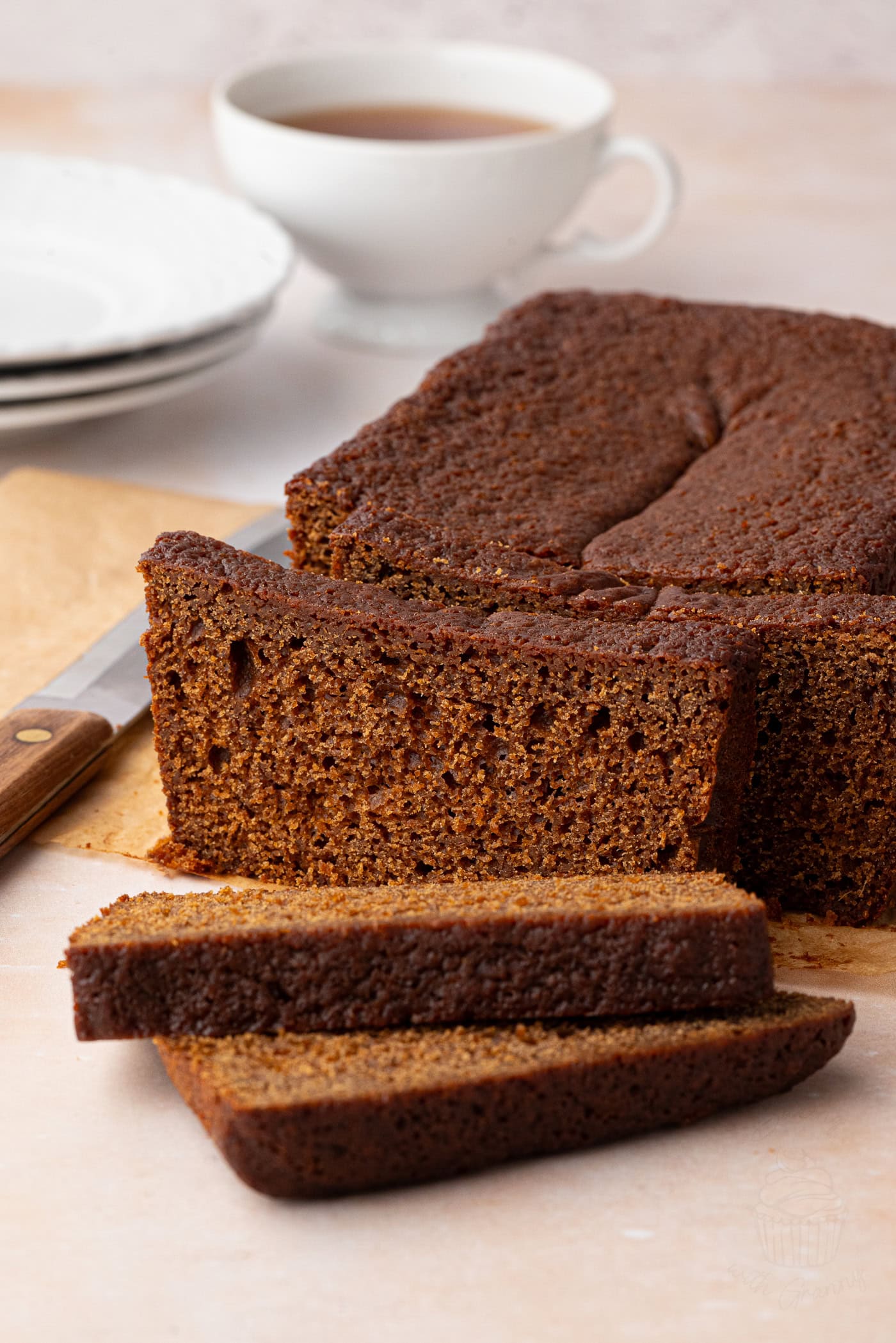 A loaf of gingerbread loaf cake rests enticingly on a wooden board, partially sliced with two pieces in the foreground. A knife lies beside it, ready for more. In the background, a cup of tea and stacked plates sit on a light-coloured surface, completing the cosy scene.