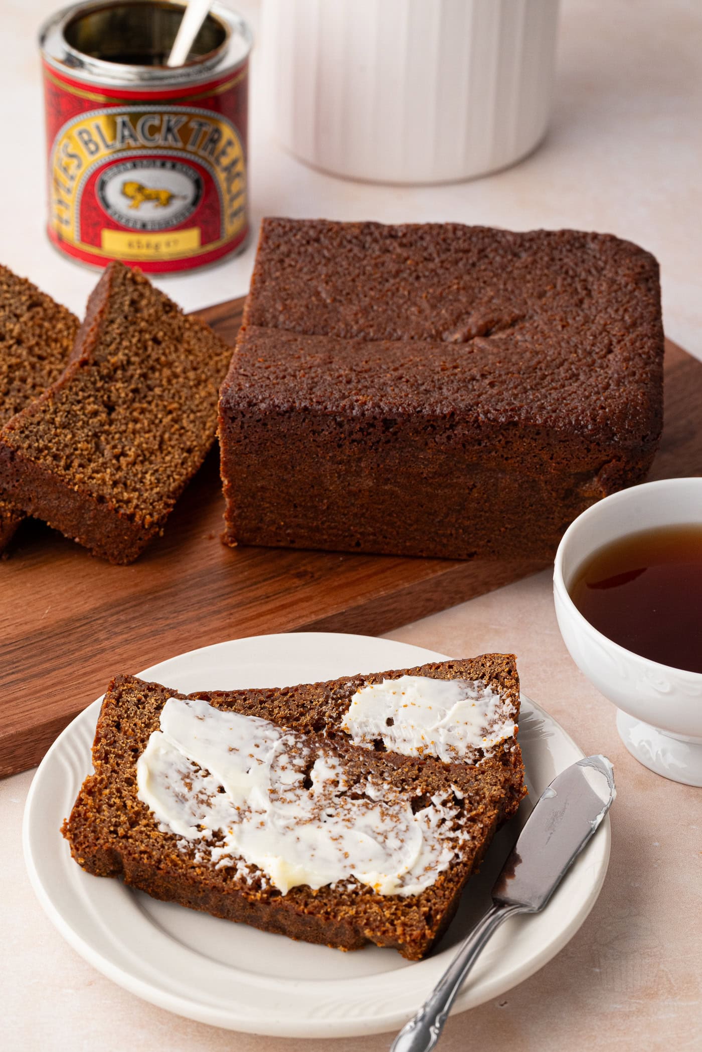 A sliced loaf of dark gingerbread loaf rests on a wooden board, with two slices on a plate. One slice is buttered, accompanied by a butter knife. A white cup with tea sits beside the plate. In the background, a tin of treacle stands next to a white jar.