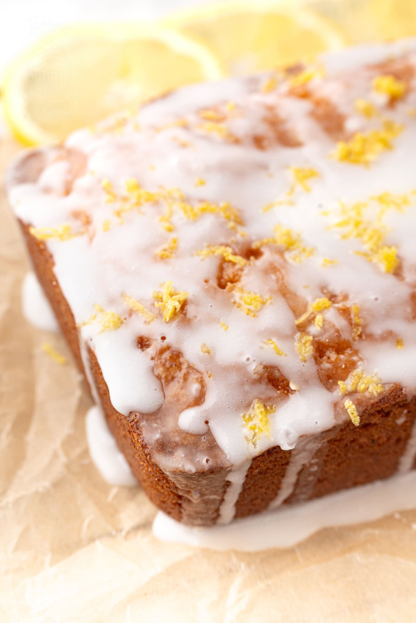 A close-up of a glazed lemon drizzle loaf cake on parchment paper. The cake is topped with a white icing and sprinkled with lemon zest. Slices of lemon are visible in the background.