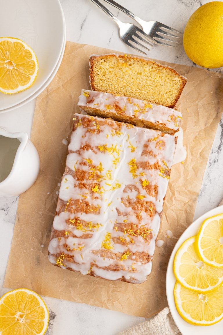 A lemon drizzle loaf cake with icing and lemon zest on top sits on parchment paper, embodying the perfect recipe. Slices of lemon surround the cake. Nearby are a pitcher, two forks, plates, and a whole lemon on a marble surface.