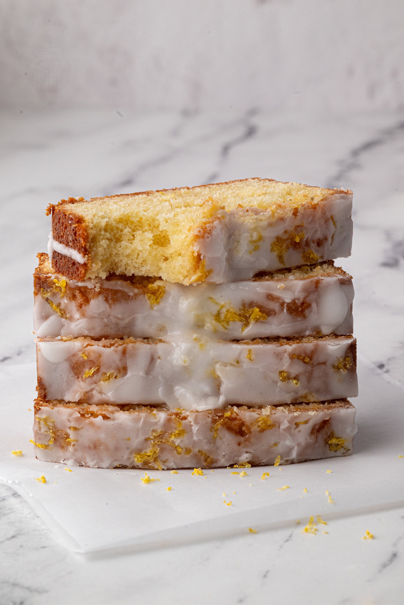 A stack of four slices of lemon drizzle loaf with white icing sits on a marble surface. The icing glistens, and small bits of lemon zest are visible on top, adding texture and colour to the golden-brown cake.