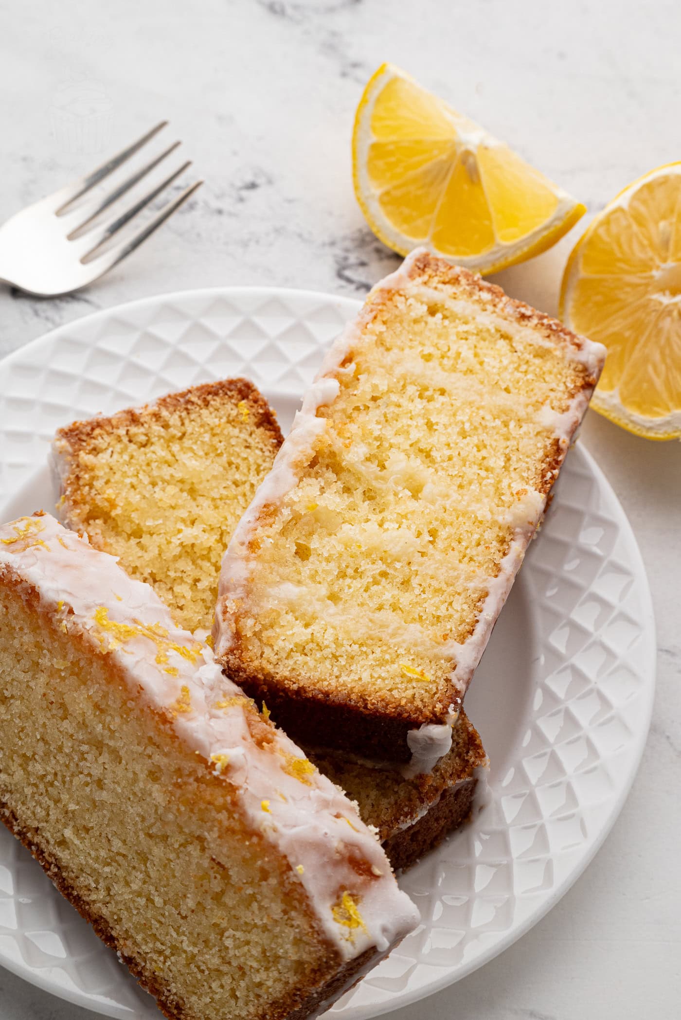 Slices of lemon drizzle loaf cake with icing and zest are served on a white plate. Two lemon wedges accompany the plate, while a fork rests on the marble surface nearby.