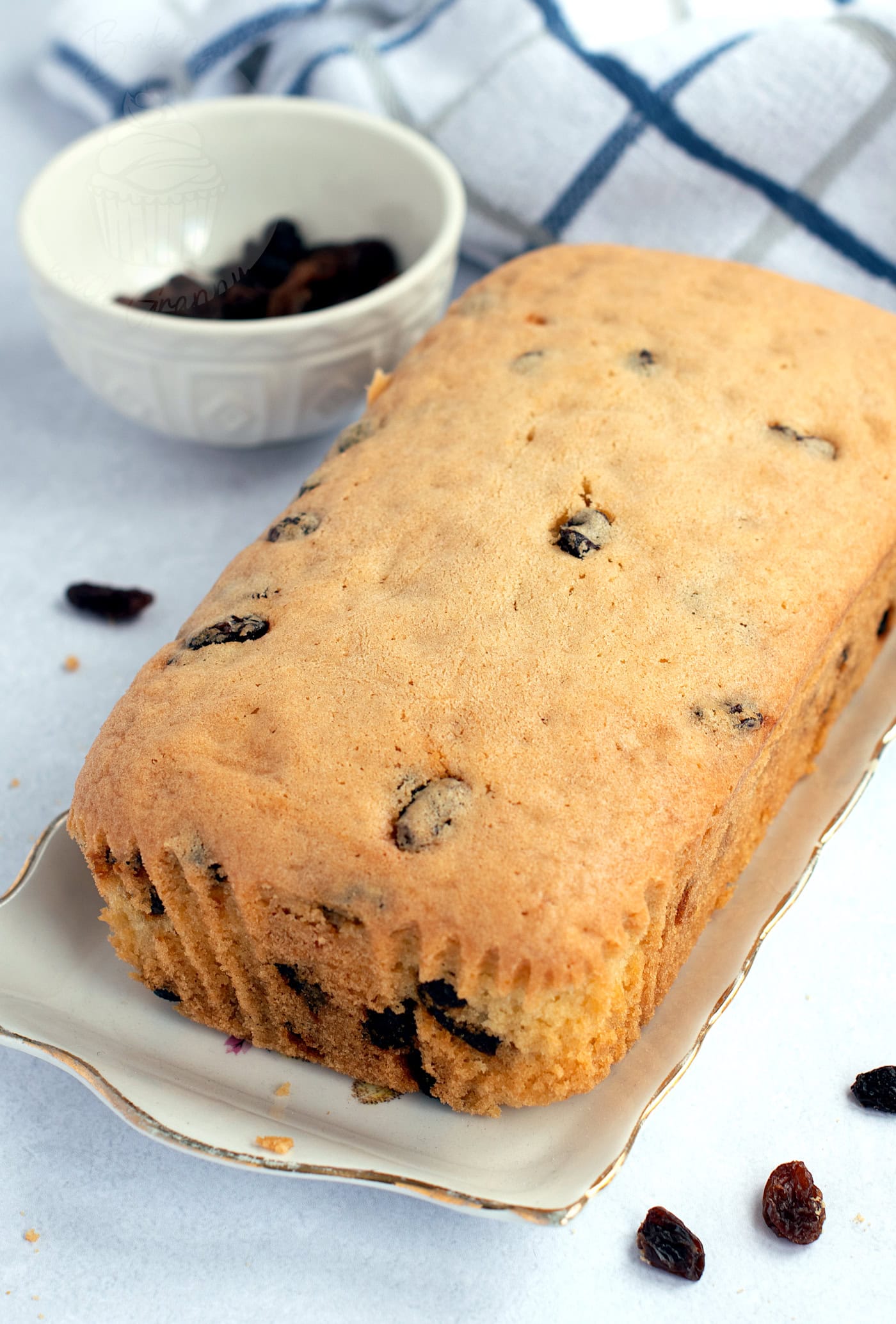 A freshly baked sultana cake rests on a rectangular platter, with a few sultanas scattered around. A white bowl brimming with more sultanas sits in the background, all elegantly arranged on a white surface adorned with a blue and white checked cloth.