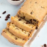 A loaf of Sultana sits on a white platter with three slices cut. Set on a light blue surface and scattered with sultana, a small white bowl of sultanas and part of a checked cloth add charm in the background.