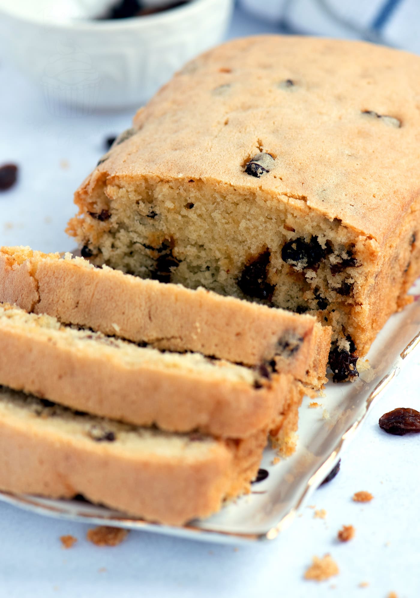 A loaf of sultana cake sits on a white rectangular plate, with several slices invitingly cut. The cake boasts a golden-brown crust dotted with juicy sultanas. Crumbs are scattered around, and a small bowl peeks into view in the background.
