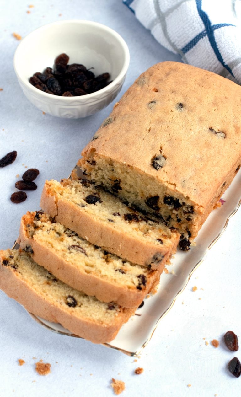 A loaf of Scottish sultana cake recipe, sits partially sliced on a plate, accompanied by a small bowl of sultanas. In the background, a checked cloth rests on the light surface.