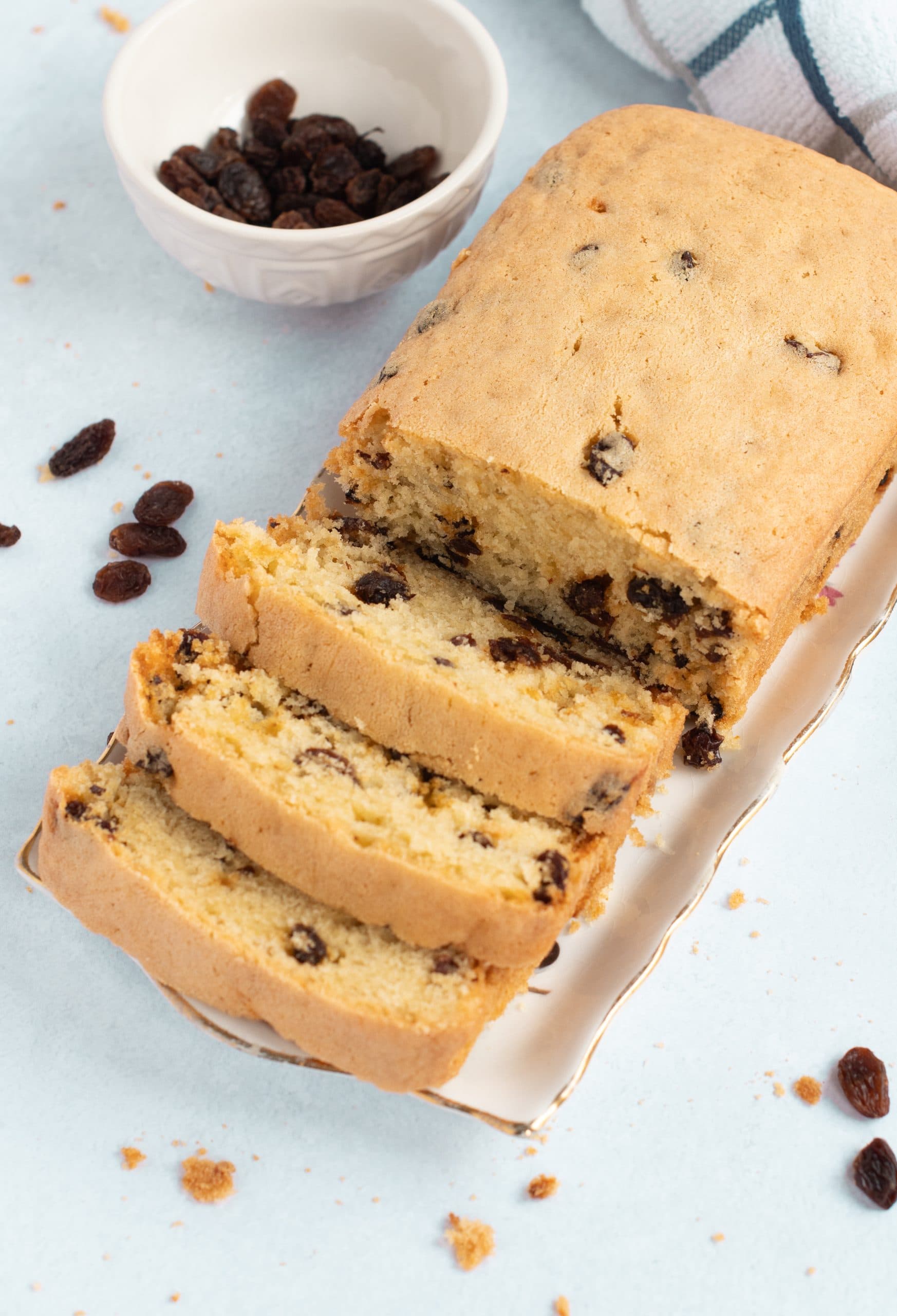 A loaf of Sultana sits on a white platter with three slices cut. Set on a light blue surface and scattered with sultana, a small white bowl of sultanas and part of a checked cloth add charm in the background.