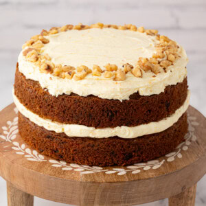 Two-layer carrot cake topped with buttercream and chopped walnuts, displayed on a wooden cake stand with inlaid detail.