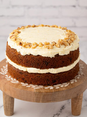 Two-layer carrot cake topped with buttercream and chopped walnuts, displayed on a wooden cake stand with inlaid detail.