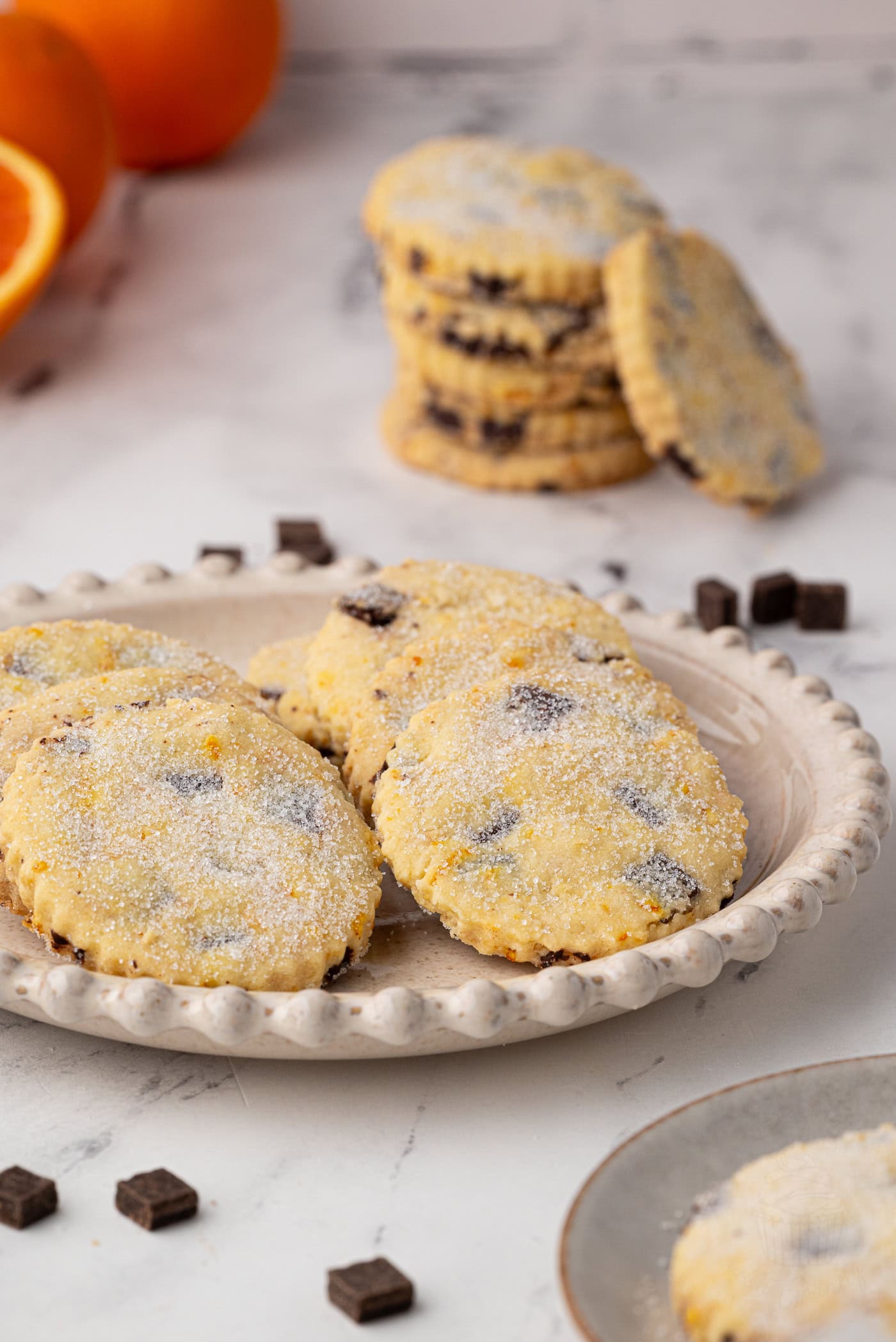Plate of chocolate orange shortbread biscuits in the foreground, with a stack of biscuits and fresh oranges in the background.