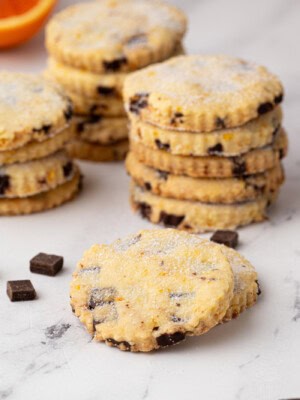 Focus on a single sugared chocolate orange shortbread biscuit, with several stacks of biscuits and an orange half in soft focus behind.