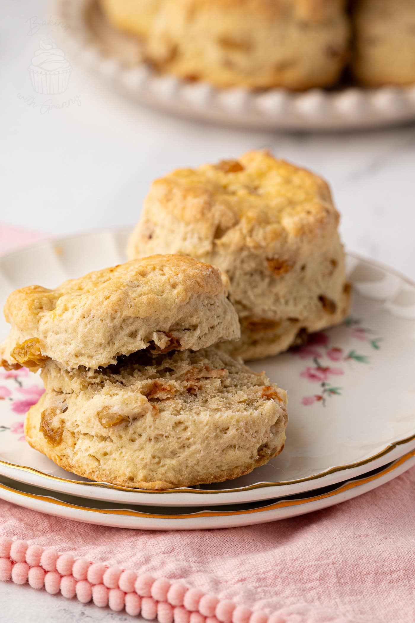 Pair of golden Fruit Scones on a vintage plate, with a batch of scones visible in the background.