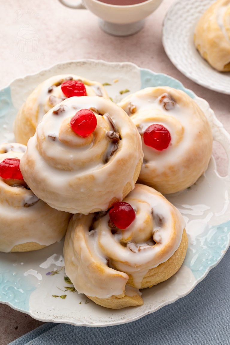 Stack of glazed Belgian buns with red cherries on top, served on a patterned plate, ideal for British teatime.