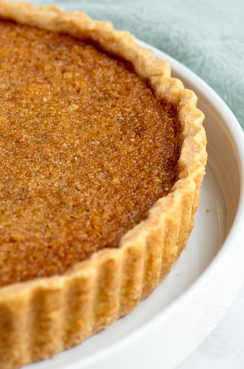 Golden treacle tart in a white dish, showing the deep amber filling and flaky shortcrust pastry edges. A classic UK dessert.