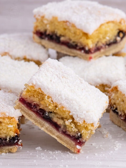 Scottish Snow Cake slices stacked on a white surface, showing the jam and sponge layers beneath a dusting of coconut.