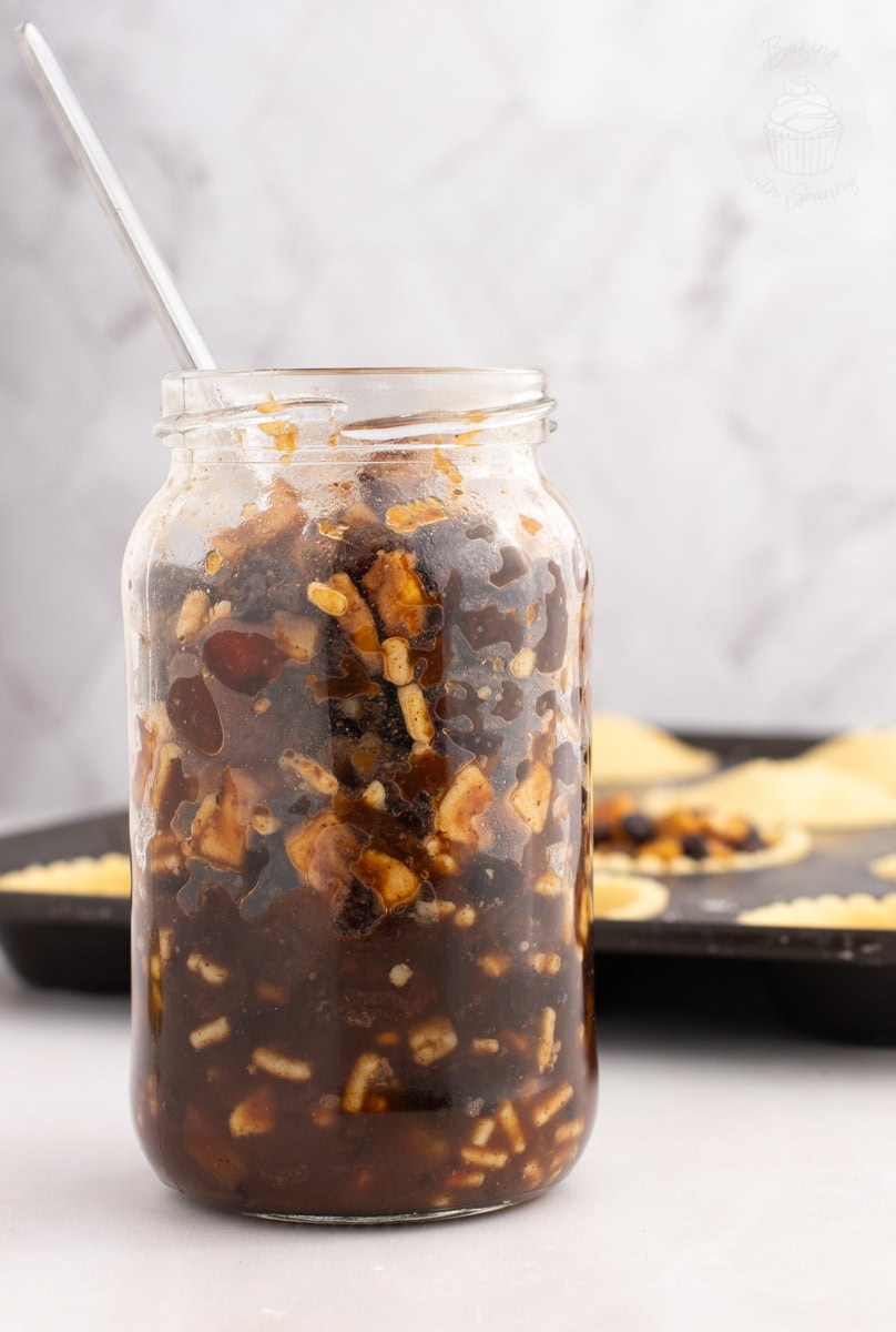 Glass jar filled with homemade British Christmas mincemeat, with a spoon inside and a tray of mince pies in the background.